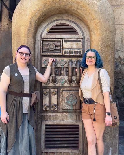 The image features two women standing side-by-side in front of an ornate metal door, likely from the Star Wars franchise.

On the left stands a woman with dark hair styled in braids and wearing a gray vest over a white shirt. She has her right hand on the door handle, which is shaped like a lightsaber hilt. On the right is another woman with blue hair and glasses, dressed in a white shirt, brown shorts, and a tan vest.

The background of the image appears to be a stone wall, possibly part of a building or structure from the Star Wars universe. The overall atmosphere suggests that the women are posing for a photo opportunity within this themed environment.