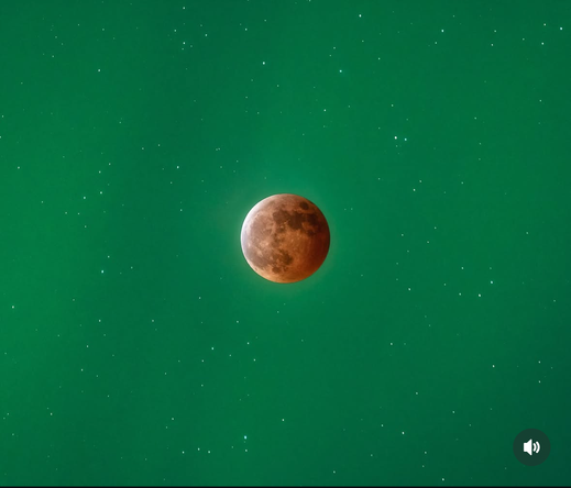 A closeup photo of the fully eclipsed moon. It’s got a reddish hue because of the earthshine. The background of stars is an ocean of green! This is because the aurora / northern lights were also happening at the same time.