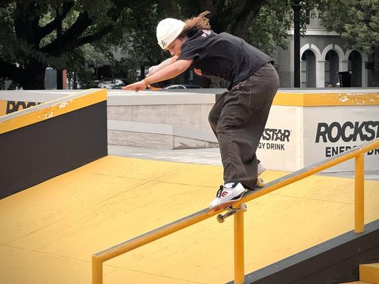 A female skateboarder performs a backside feeble grind down a handrail on a mostly cloudy day.