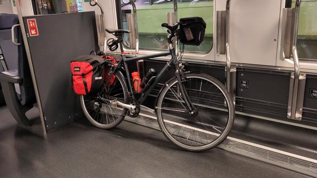 Un vélo gris avec ses sacoches rouges est attaché dans le compartiment vélo du train.