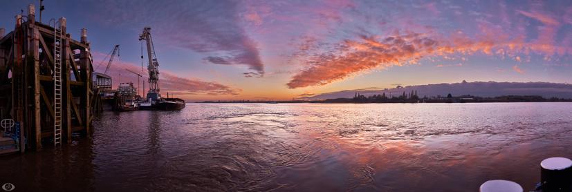 The river scheldt during sunset. Sun has already gone under putting the clouds in vibrant (orange) colours. At the left bank a pier and a little further ahead industry with chimneys and cranes. At the right the right bank of the river where a nature reserve begins.