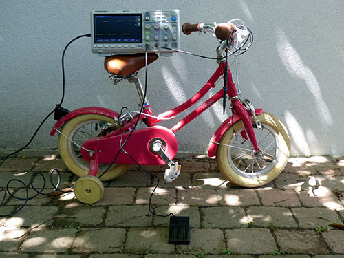 Un oscilloscope posé sur la selle d'un vélo rouge pour enfant (avec les roulettes).
Une pédale noire est posée devant branchée au système. Une sonde de l'oscilloscope est enroulée au guidon et un signal carré apparaît en jaune sur l'écran de l'oscilloscope