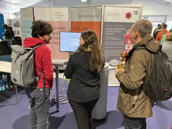 Gesprächssituation am Stand der TIB. Eine TIB-Mitarbeiterin zeigt einem interessierten Tagungsteilnehmer stehend am Computerbildschirm den ORKG. Ein weiterer Man steht interessiert zuhörend daneben. Im Hintergrund vier Rollup-Banner mit TIB-Services.