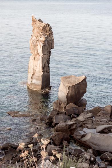 Two column-shaped rocks emerge from the sea near a cliff. The sea is calm and the light is warm, early in the morning.