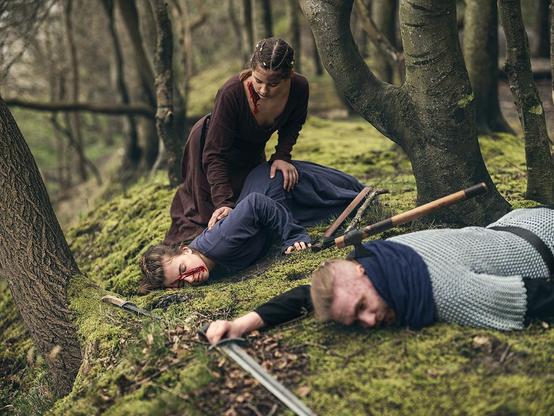 Ein Wikingermädchen kümmert sich um ein anderes, verletztes Mädchen, das vor ihr auf dem Waldboden liegt. Im Vordergrund liegt ein erwachsener Wikinger mit Axt im Rücken
