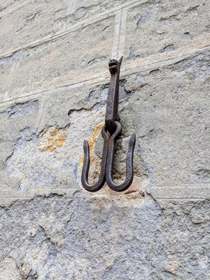 A close-up shot shows a weathered, dark metal hook affixed to a rough, light grey stone wall. The hook has a decorative top resembling a stylised animal head, and below it, two curved prongs extend downwards. The metal appears aged and slightly rusted. The stone wall has visible mortar lines and variations in texture and colour, indicating its age and exposure to the elements.