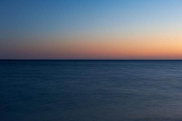 The photo shows the sea and the sky after the sunset, during blue hour. The horizon is exactly in the middle. The lower part displays a milky dark blue sea due to the long exposure. The upper part shows from the horizon up a finely graduated transition from orange to light blue of the sky.