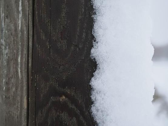 This image shows a close-up of a wooden surface with a layer of snow on it. The wood appears weathered, and the snow has a textured, crystalline appearance.