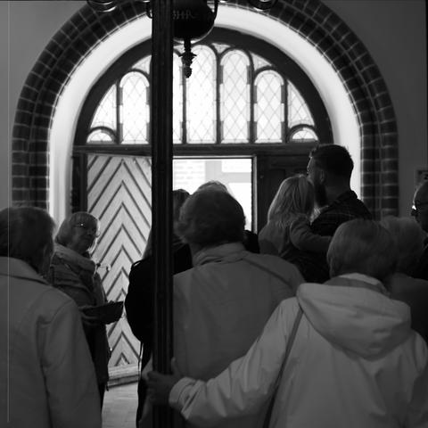 This black and white image shows a group of people gathered in front of an ornate door with stained glass windows. The scene appears to be indoors, possibly in a church. The atmosphere seems to be calm and communal.