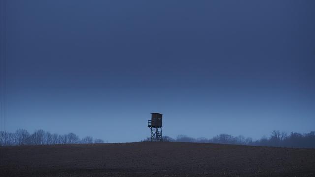 The image depicts a solitary wooden watchtower or hunting blind standing on a hill against a backdrop of a dark, overcast sky. The landscape appears barren and the horizon features silhouettes of trees. The overall mood is calm and atmospheric.