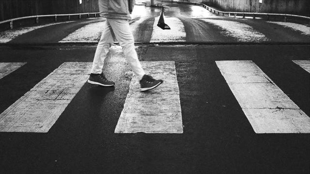 Das Schwarzweißfoto zeigt einen Zebrastreifen von der Seite während eine Person diesen von links nach rechts überquert. Der Oberkörper der Person ist außerhalb des Fotos. Sie trägt eine helle Hose und dunkle Schuhe mit hellen Sohlen. Im Hintergrund deuten Betonwände, Geländer und ein Gefälle auf eine Tiefgarageneinfahrt hin. Leichter Schneefall und Fahrzeugreifen haben ein Muster erzeugt, das dem Zebrastreifen ähnelt.

The black and white photo shows a crosswalk from the side as a person crosses it from left to right. The person's upper body is outside the photo. They are wearing light-colored trousers and dark shoes with light-colored soles. In the background, concrete walls, railings and a slope indicate an underground parking garage entrance. Light snowfall and vehicle tires have created a pattern that resembles the crosswalk.