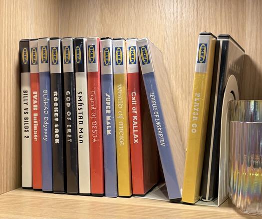 A stack of video game boxes on a shelf with IKEA related versions as titles:
PLATSA GO
LEAGUE OF LAGKAPTEN
CALL OF KALLAX
WORLD OF MICKE
SUPER MALM
LEGEND OF BESTÅ
SMASTAD Man
FEKET
ROCKET LACK
BLÀHAJ Odyssey
IVAR Infinate
BILLY VS BILDS 2