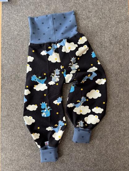 Auf einer grauen Unterlage liegt eine Baby-Pumphose. Der Stoff ist dunkelblau mit kleinen hellblauen Drachen, kleinen gelben Sternen und weißen Wolken. Das Bündchen an Bauch und Beinen ist mittelblau mit dunkelblauen Sternen.
