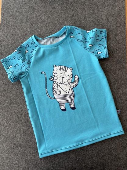 Auf einer grauen Unterlage liegt ein hellblaues T-Shirt mit einer schwarz-weiß gestreiften Katze. Die Katze ist eher Comic-Artig. Steht aufrecht, hat ein Halstuch um und hebt die eine Pfote, als würde sie winken.
Auf den Ärmeln sind Dreiecke mit dunkelblauem Rand und kleinen Punkten.