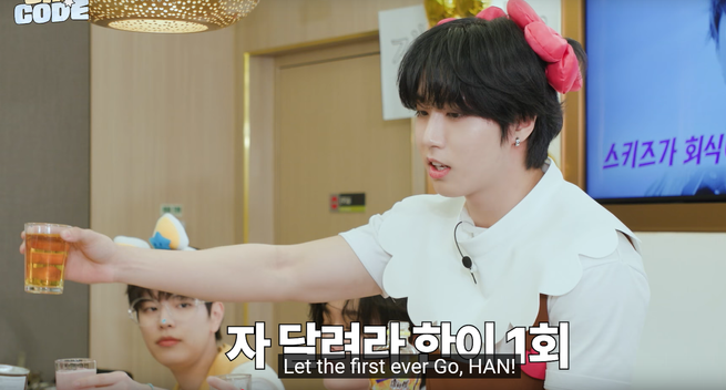 Han proposes a toast. Seungmin holds up a glass of something pink and fizzy. Caption: Let the first ever Go, HAN!