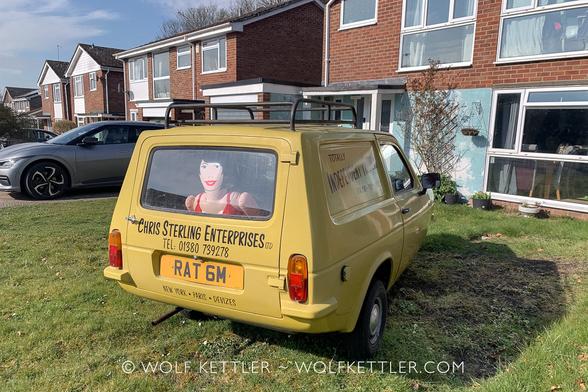 A yellow three wheeled Reliant Regal parked outside a row of dated looking domestic properties. Written on the back is “Chris Sterling Enterprises”. The registration number is RAT 6M. Looking out the back window is a female blowup doll.