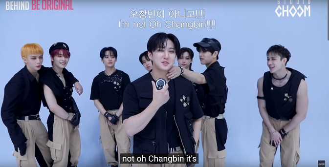 Stray Kids at Studio Choom filming some content where they're supposed to estimate time using a stopwatch. Changbin is closest to the camera, holding up the stopwatch and trying to concentrate. Seungmin stands behind him and reaches out to caress the side of his neck. Everyone else is watching extremely attentively. Caption: I'm not Oh Changbin!!!!
