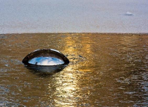 A metal lamp in a frozen pond. At the top a horizontal stripe, about a third of the photo is frozen water, white because covered with soft snow. The bottom two thirds are ice wet by a fountain and colored gold by the sun. On the left in the middle half of a shiny metal round lamp. The blue sky reflects in it. Along the lamp vertically a bright golden stripe of sunlight.