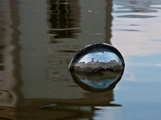 The same lamp, but now right in the middle. Right a wide vertical stripe of blue sky reflected in the water. Left the reflection of the building behind the pond. The blue sky reflects in the metal lamp. The lamp itself also reflects in the water, making it look like a round blue ball.