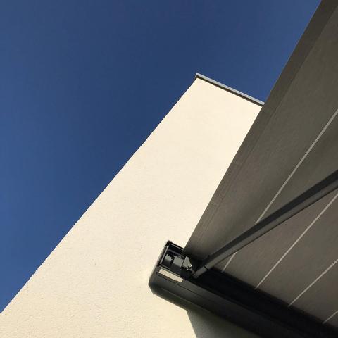 Das Bild zeigt einen Teil eines modernen Gebäudes in einem schrägen Blickwinkel. Die Fassade des Gebäudes ist weiß und glatt. An der rechten Seite befindet sich eine graue, horizontal gestreiften Markise, die nur teilweise zu sehen ist. Der Himmel ist klar und tiefblau, was auf einen sonnigen Tag hinweist.
