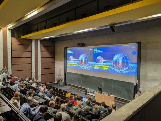 Blick in einen gefüllten, großen Uni-Hörsaal mit Doppelprojektion einer Präsentationsfolie zur Buchvorstellung.