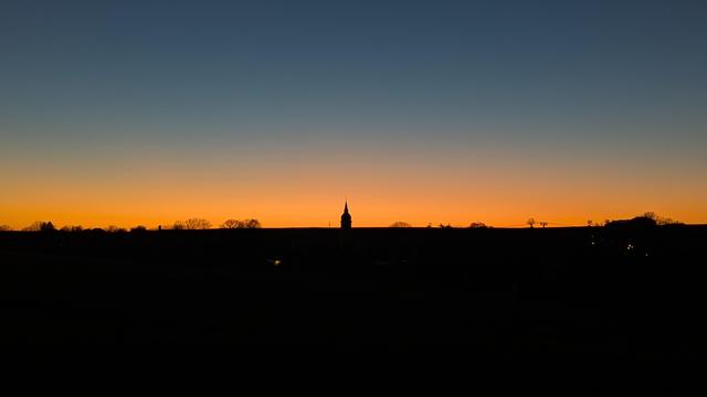 Cloudless sunset with black landscape contour in the bottom half and a color blend from orange in the middle to a blue-grey sky in the top.