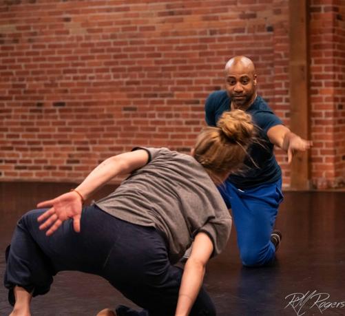 A photograph of a man and a woman participating in a modern dance class. They are in casual loose-fitting clothing, kneeling on the floor.