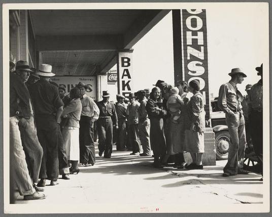 The image depicts a black and white scene with numerous people standing in line on a sidewalk, suggesting an organized event or gathering. They are waiting patiently against the backdrop of what appears to be storefronts; one prominently displays "Bakery" above its entrance. Many individuals wear hats, indicating possible sunny weather or as protection from elements. The group is diverse, including men, women, and children in various attire that suggests a bygone era, possibly mid-20th century America.

Prominent signage includes the words "Johnson Bakeries," alongside recognizable logos of Coca-Cola and another brand with partial visibility due to cropping. A vintage car's tire hints at an earlier automotive design. The overall atmosphere is one of anticipation or routine procedure, as these people await their turn for a service, likely in connection with financial aid.

The image exudes historical context, hinting at economic conditions that necessitated organized relief checks and the societal impact on individuals' daily lives during this period.