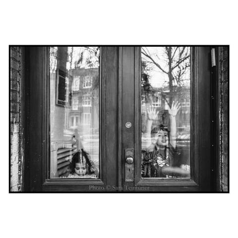 (English follows) Derrière une porte vitrée, deux enfants se tiennent. L'un regarde l'objectif photographique, tenant les bras au-dessus de sa tête et appuyés sur la vitre. L'autre, plus jeune, de l'autre bord de la porte vitrée, ne dépasse que de sa tête et regarde vers le bas. Sur les vitres, se reflètent les bâtiments et les arbres de la rue. 

Behind a glass door, two children stand. One is looking into the camera lens, holding his arms above his head and leaning against the glass. The other and younger child, on the other side of the glass door, does not show more than her head. She is looking down. The buildings and trees of the street are reflected on the glass.