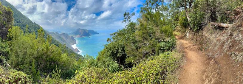 Panorama-Aufnahme von einem erdigen Fusspfad auf der rechten Seite, der an einer Steilküste zwischen Felsen, Büschen und Bäumen in der Mitte des Bildes den Blick in die Ferne auf das hellblaue Meer mit weißer Gischt an der Inselküste und den weiß-bewölkten, blauen Himmel freigibt.