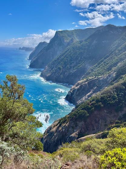 Blick von einer Fels-Steilküste auf das blaue Meer und den blauen Himmel mit wenigen weißen Wolken. Rechts kann man die nordwestliche Steilküste Madeiras entlang schauen.