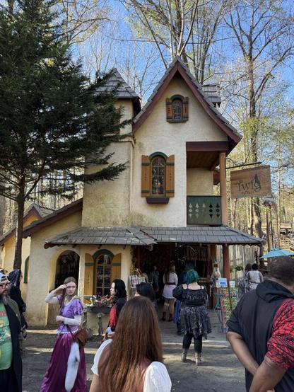 A whimsical, fairy-tale-style shop at a Renaissance fair with people in costumes browsing and walking past under a bright blue sky.