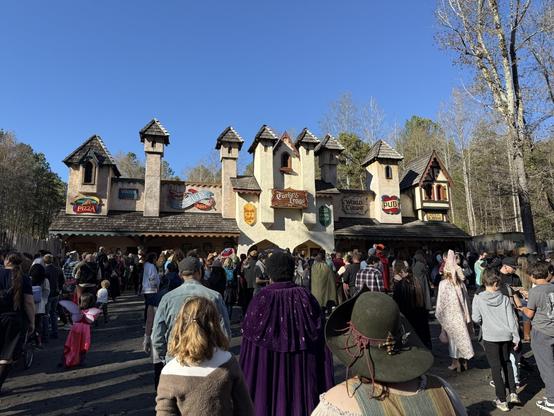 A bustling food court at a Renaissance fair with a medieval-style facade, serving pizza, soup, turkey legs, and more, as costumed guests gather.