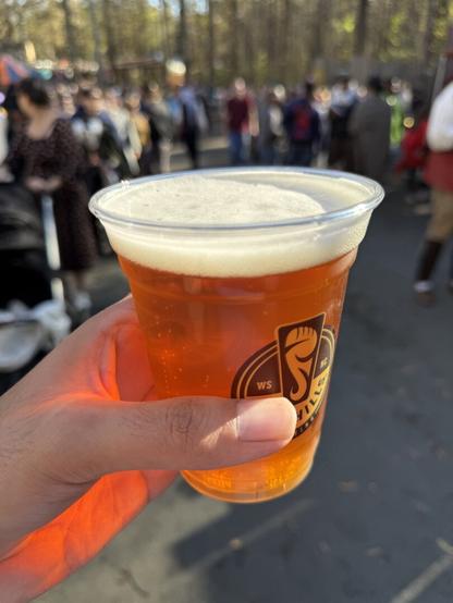A hand holds a plastic cup of pumpkin ale with a frothy head at a Renaissance fair, with a blurred crowd in the background.