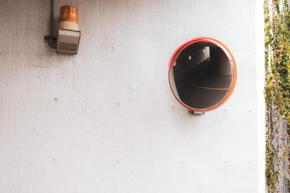 The image shows a wall with an orange warning light fixture and a convex mirror. The mirror is typically used for surveillance or to provide visibility around corners. There are some plants climbing on the wall's edge.