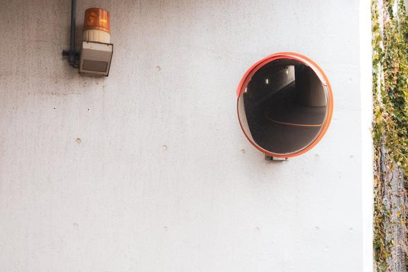 The image shows a wall with an orange warning light fixture and a convex mirror. The mirror is typically used for surveillance or to provide visibility around corners. There are some plants climbing on the wall's edge.