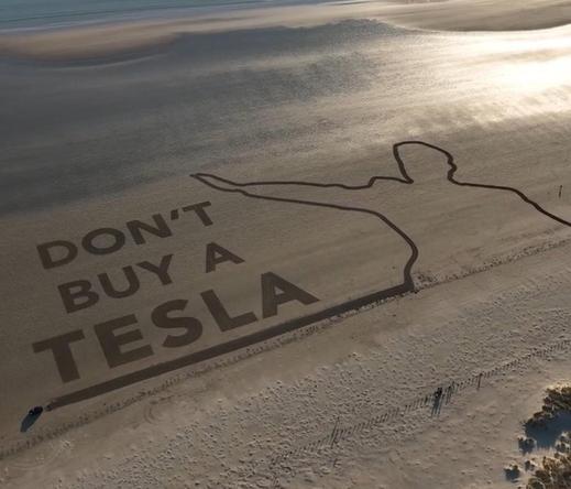This image shows a large sand drawing on a beach with the text "DON’T BUY A TESLA" along with a silhouette of a Elon Musk doing the Hitler salut. The location is Black Rock Sands, Wales, UK, and the artwork was created by Led By Donkeys, a UK-based activist group known for their large-scale public messages, often targeting political and corporate issues.