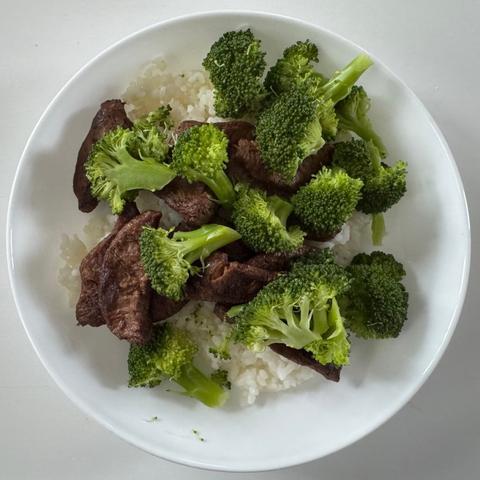 A white shallow bowl filled with white rice, dark brown faux meat, and bright green broccoli crowns