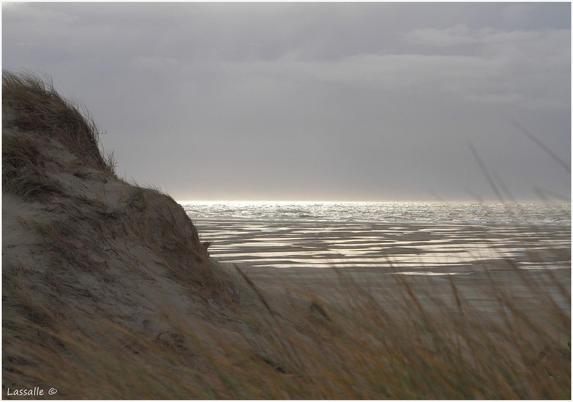 Der Blick durch einen schmalen Dünenübergang zum Strand. Links der mit Seegras bewachsene Dünenhang, rechts der Blick durch einige Seegraspflanzungen. Das Wasser wird durch die schon tiefer stehende Sonne beschienen und reflektiert, besonders am Horizont, besonders grell. Der Himmel stark bewölkt bis grau, nur eine kleine Lücke bleibt, um diesen Lichteffekt zu erzeugen.