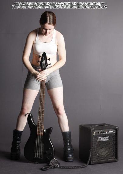A white woman wearing black books and grey pants holding a black bassguitar by the neck/head, the bass guitar standing upright on the ground, her legs spread on either side of the bass. The bass is plugged into an amp.