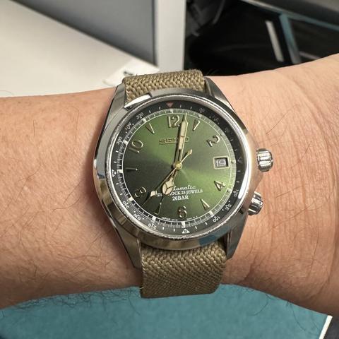 Watch with brushed and polished stainless steel case, internal rotating compass bezel, cathedral hands, sunburst green dial with 3:00 date, gold numeric indices at 12, 2, 4, 6, 8, and 10, gold dagger indices at 1, 5, 7, 9, and 11, and dual crowns at 3:00 and 4:00. On a wheat-colored woven fabric strap.