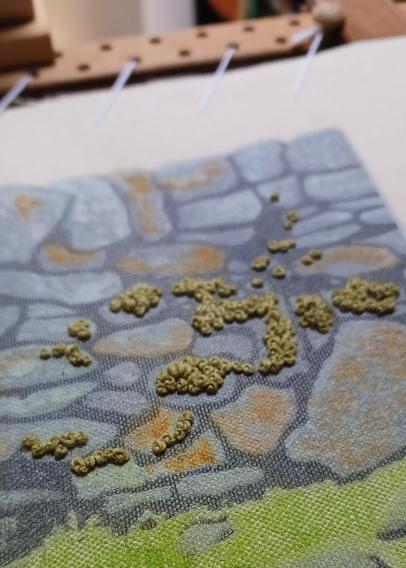 A linocut print on fabric depicting a dry stone wall. Stitches in green wool resemble moss growing on the wall. The fabric is stretched on an embroidery frame and tilted to dhow texture