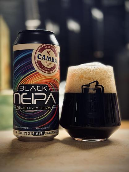 A can of beer with a rainbow swirl and the words ‘Camba Black NEIPA New England IPA’ sitting next to a full glass with large foamy head and dark, opaque beer