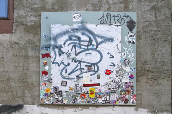 As described in the post, a metal plate bolted to a cement wall in an alley.  It's been there years and is weathered but still continues to collect graffiti in spray paint, marker or chalk, with stickers at random filling the bottom and partway up the sides of the square plate painted light gray-green. The outer areas are dirty textured cement, gray with brown details in cracks.  A bit of a window is top left but looks sealed up. Grungy image but this is an actual photo not edited with filters.