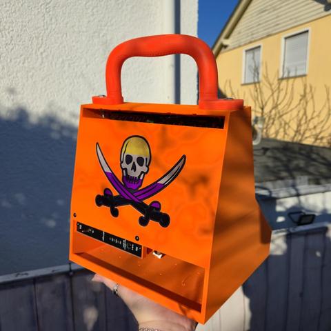 A bright orange, 3D-printed portable Eurorack studio case with a sturdy handle is held in one hand. The back panel features a custom pirate sticker with a skull and crossed swords in non-binary colors. Below the sticker, the case has the power input port. There is a big opening on the lower part and a small on top of the back(airflow) The case is photographed outdoors in sunlight, with a white wall and a house with closed shutters in the background.
