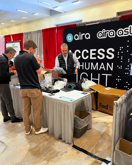 Aira team members set up a booth at CSUN 2025, unpacking boxes and organizing materials. A black banner behind them displays the Aira logo and the phrase “ACCESS IS A HUMAN RIGHT.” The booth is surrounded by red and gray curtains.