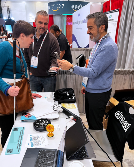 A woman with a white cane interacts with two Aira team members at the booth. One holds a small device while the other shows something on a smartphone. The table is covered with flyers, tech accessories, and a laptop. A black Aira ASL shirt is draped over a chair.