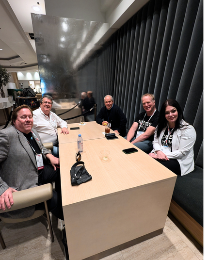 Five people sit around a wooden table in a lounge area, engaged in conversation. Two wear black “Access” shirts, while others wear business attire. A reflective silver divider and dark gray cushioned walls are in the background.
