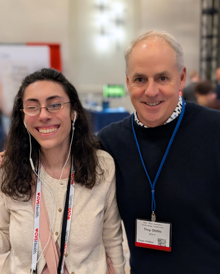 A smiling woman with glasses and earbuds stands next to a man wearing a navy sweater and a blue lanyard labeled “Troy Otillio, Aira.io, Expo Visitor” at CSUN 2025. The background shows a blurred conference setting with booths and people.