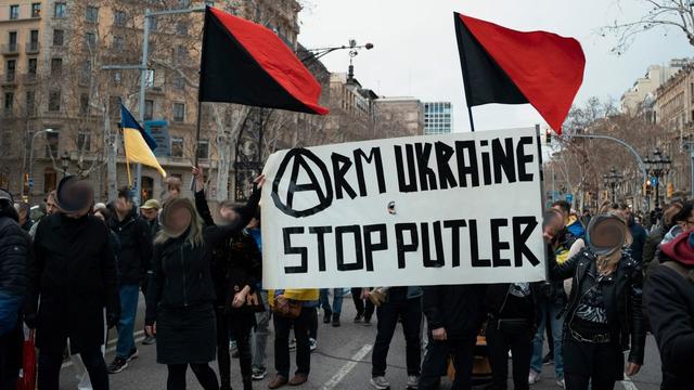 Picture of a demonstartion with a banner saying: (A)RM UKRAINE STOP PUTLER . 3 flags held by demonstrators, 2 are anarcho-communist red and black flags and 1 is the Ukrainian blue and yellow flag.